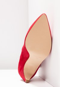 Chaussure à talon haut pointu en daim rouge reposant avec la semelle tournée vers l'avant, taille 37, fabriquée en Espagne, sur un fond d'angle blanc.