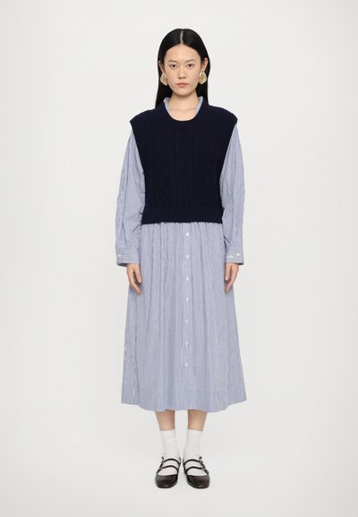 Granatowy sweter bez rękawów nałożony na niebieską i białą w paski sukienkę koszulową. Sukienka ma długie rękawy i długość do połowy łydki, zestawiona z czarnymi butami.