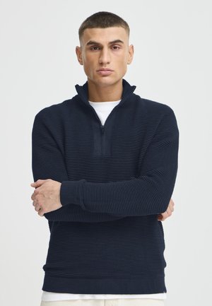 Homme aux cheveux courts portant un pull marine côtelé à fermeture éclair quart, par-dessus une chemise blanche, debout les bras croisés devant un fond uni.