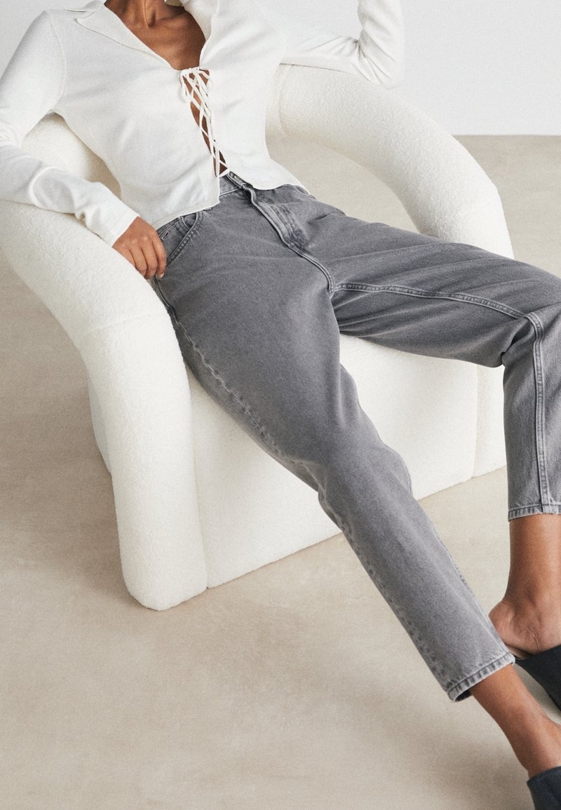 Una persona se sienta en una silla blanca y lujosa, vestida con una blusa blanca con cordones y jeans grises, en un espacio interior minimista.