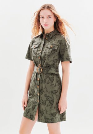 Robe chemise vert olive à manches courtes, devant à boutons, deux poches poitrine et une ceinture à la taille avec une boucle ronde. Motif floral sur l'ensemble.