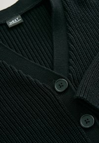 Zwarte gebreide cardi met een V-hals, geribbelde textuur en twee zwarte knopen aan de voorkant. Label zichtbaar bij de halslijn.
