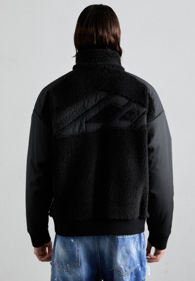 Schwarze Jacke mit einem strukturierten, fluffigen Rückenteil und glattem Vorderelement, ausgestattet mit einem hohen Kragen und einem Logodetail über den Rücken.