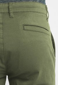 Pantaloni in cotone verdi che mostrano una tasca posteriore piatta, dettagli cuciti e una texture liscia, con una cintura visibile sopra.
