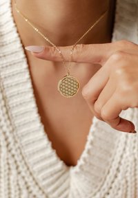 Collar de oro con un patrón geométrico, suspendido de una delicada cadena, sostenido entre los dedos, usado sobre un suéter blanco texturizado.