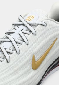 Krupni plan bijele sportske tenisice s teksturiranom tkaninom, sivo-bijelim vezicama i zlatnim Nike swoosh logom s strane.