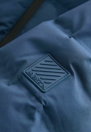 Nahaufnahme eines marineblauen Stoffes mit einem erhabenen quadratischen Patch, der diagonale Linien und den embosst eingeprägten Text "40 26'72'' N" zeigt.