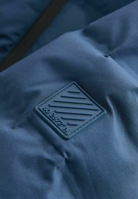 Gros plan d'un tissu matelassé bleu foncé avec un patch carré en caoutchouc surélevé montrant des lignes diagonales et les coordonnées "40.2672°N."