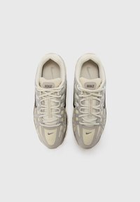 Sneakers Nike z jasnobeżową i szaro-materiałową cholewką, teksturowanymi nakładkami i grubą gumową podeszwą. Widoczne są sznurowadła oraz logo.