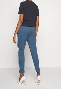 Osoba w niebieskich, teksturowanych spodniach z białymi paskami po bokach, czarnej koszulce z krótkim rękawem i białych szpilkach, stojąca przy białej ścianie.