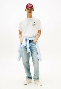 Witte katoenen T-shirt met kleurrijk "LMC" logo, lichtblauw en wit gestreept shirt om de taille geknoopt, lichtblauwe jeans, beige sneakers, rode pet.