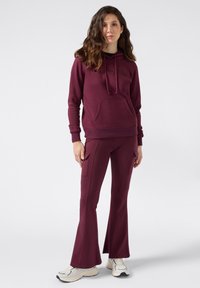 Sudadera burdeos con cordón y bolsillo frontal tipo canguro, combinada con pantalones acampanados a juego que presentan bolsillos laterales tipo cargo. Zapatillas blancas completan el conjunto.