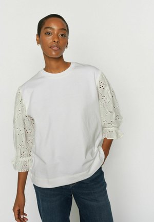 Frau mit kurzen Haaren, die ein weißes Oberteil mit Lochspitzen-Puffärmeln und dunkelblaue Jeans trägt, vor einem schlichten, hellen Hintergrund stehend.