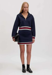 Navy-blaues Strickkleid mit einem Reißverschlusskragen, der rote und weiße horizontale Streifen an der Taille und an den Ärmeln aufweist. Kombiniert mit einer schwarzen Clutch.