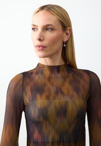 Langsleeve, doorschijnend top in een multicolore stof met abstract patroon, met een hoge hals en ribbelstructuur. Kleine stervormige oorbellen.