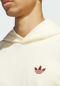 Cream hoodie with a drawstring, featuring a red Adidas logo on the chest. Soft texture, fitted design, and no visible patterns.