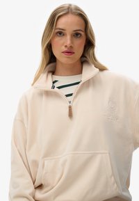 Pullover in pile beige con colletto alto e zip a metà. Presenta una tasca frontale e un dettaglio del logo ricamato sul petto. Texture liscia.
