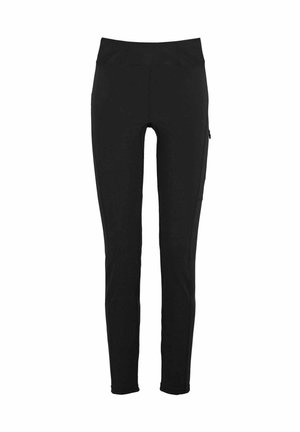 Vrouw met lang gevlochten haar, staand en naar de camera kijkend, draagt een beige shirt met korte mouwen, zwarte leggings met zakken, zwarte schoenen en sokken.