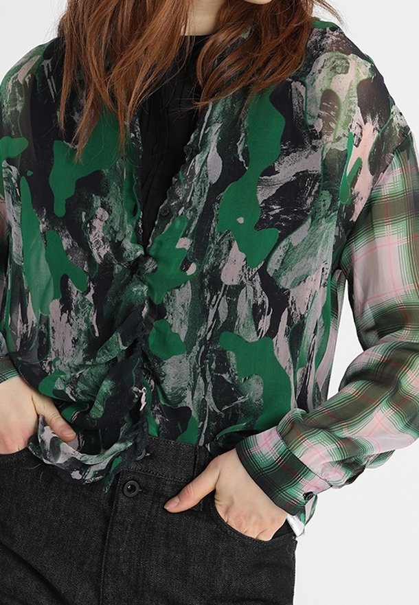 Ma na sobie zieloną, czarną i różową bluzkę w abstrakcyjny wzór z plaidowymi rękawami, włożoną w czarne spodnie z wysokim stanem, z rękami w kieszeniach.