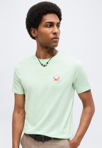 Νέος άνδρας με κοντό αφροκράτημα που φοράει ανοιχτοπράσινο μπλουζάκι με μικρό κόκκινο λογότυπο καβουριού και μαύρο κολιέ, κοιτάζοντας στο πλάι.