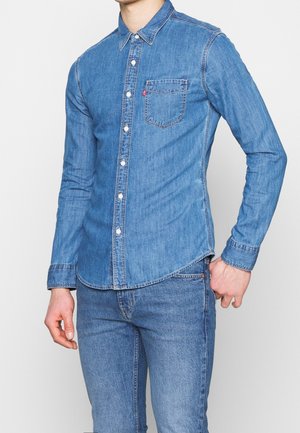Camicia di jeans blu con maniche lunghe, chiusura a bottoni, colletto a punta e taschino sul petto. Presenta una leggera curvatura sull'orlo e una texture liscia.