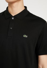 Homme portant un polo noir avec deux boutons et un petit logo crocodile vert sur la poitrine gauche.