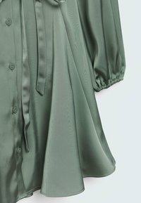 Blouse en satin vert avec un devant boutonné, des manches bouffantes et une ceinture à nouer à la taille, créant une silhouette fluide et élégante.