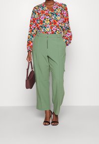 Donna che indossa pantaloni verdi a vita alta, una blusa floreale multicolore, tacchi neri e tiene in mano una piccola borsa a tracolla marrone scuro.