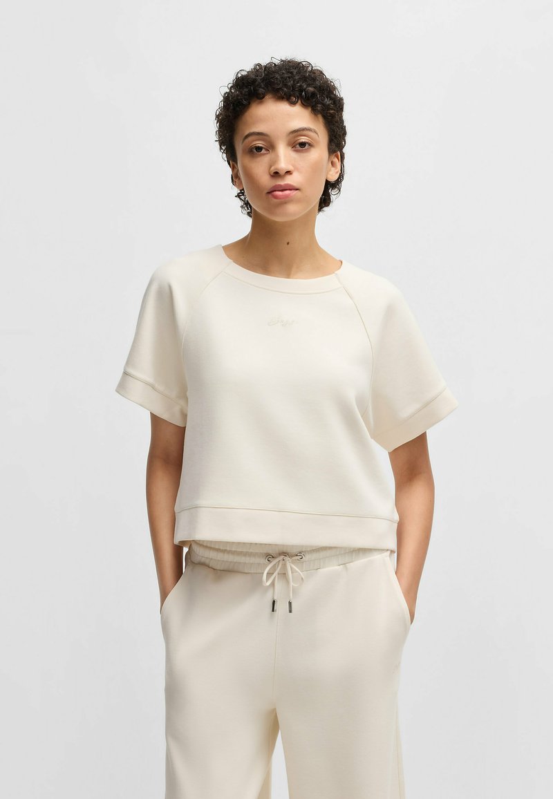 Cream-colored short-sleeve sweatshirt with a relaxed fit, rounded neckline, and subtle embroidered logo; smooth fabric with ribbed hem.