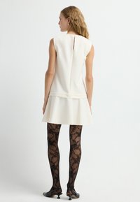 Mouwloze witte jurk met uitlopende zoom, achterkant met ritssluiting, gecombineerd met zwarte tights met kantpatroon en zwarte schoenen met hakken.