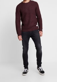 Uomo che indossa un maglione testurizzato bordeaux scuro, jeans neri a vestibilità slim e sneakers bianco e nere, in piedi contro uno sfondo bianco semplice.