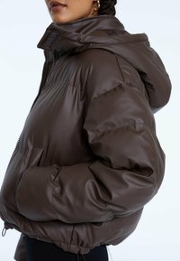 Giacca a vento imbottita marrone con una texture lucida, chiusura con zip sul fronte e cappuccio. Presenta ampie tasche laterali e polsini elasticizzati.