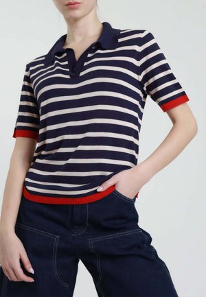 Donna che indossa una polo a righe blu navy e bianche a maniche corte con bordature rosse, abbinata a pantaloni blu scuro con cuciture visibili.