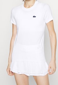 Haut blanc à manches courtes avec texture côtelée, présentant un col rond et un logo brodé bleu marine sur la poitrine gauche.