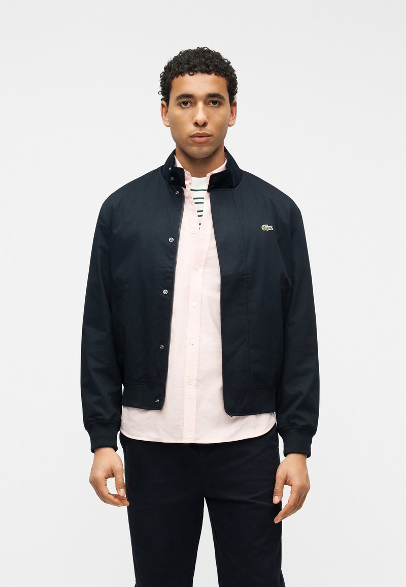 Άνδρας που φοράει ένα σκούρο μπλε μπουφάν με φερμουάρ πάνω από ένα ανοιχτό ροζ πουκάμισο με ριγέ μπλουζάκι από μέσα, στέκεται μπροστά σε λευκό φόντο.