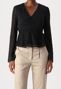 Blusa nera a maniche lunghe trasparente con pois e vita con cordoncino abbinata a pantaloni beige su una persona in piedi, con le braccia rilassate.
