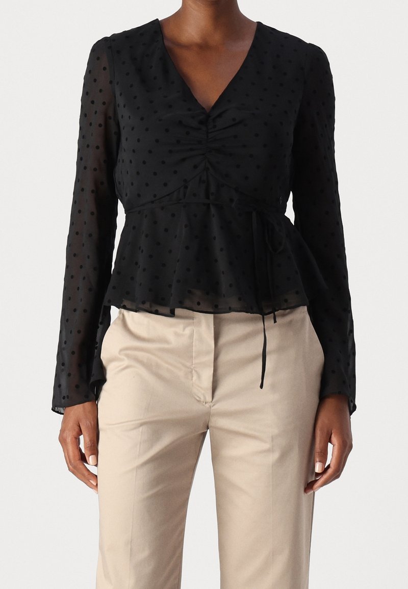 Blusa nera a maniche lunghe trasparente con pois e vita con cordoncino abbinata a pantaloni beige su una persona in piedi, con le braccia rilassate.