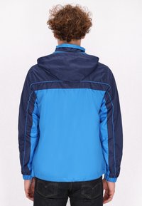 Blauwe en marineblauwe windbreakerjas met capuchon, voorzien van contrasterende panelen, elastische manchetten en lichtgewicht stof, ontworpen voor gebruik buitenshuis.