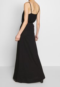 Abito maxi nero con spalline sottili, tessuto fluido e vita aderente. Presenta una texture liscia e un design minimalista, con una scollatura sulla schiena.
