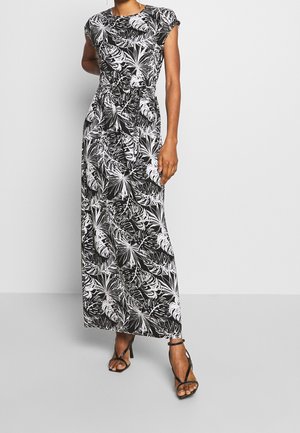 Nainen pukeutuneena mustavalkoiseen, trooppisten lehtikuvioiden maksimekkoon, jossa on lyhyet hihat, ja mustat korkokengät sandaalit, seisomassa yksiväristä seinää vasten.