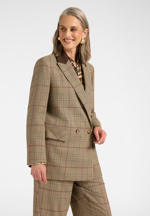 Lächelnde Frau mit grauen Haaren, die einen braunen karierten doppeltreihigen Blazer und passende Hose vor weißem Hintergrund trägt.