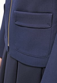 Námořnická modrá bunda s zipovým zapínáním a klopovou kapsou. Látka je strukturovaná s formálním designem, který zdůrazňuje čisté linie a minimální akcenty.