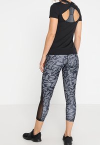 Svart atletiskt linne med öppning i ryggen, ihop med mönstrade grå leggings med meshpaneler och en figurnära passform. Svarta sneakers.