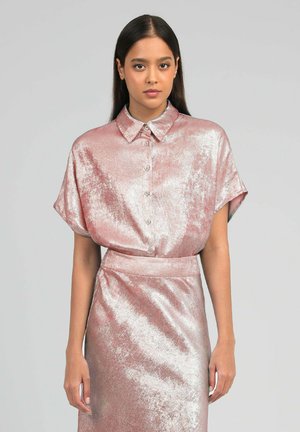 Femme aux longs cheveux foncés portant un chemisier rose texturé brillant à manches courtes boutonné et une jupe assortie sur un fond gris.