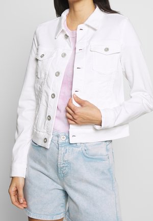 Vrouw draagt een witte denimjas met knopen over een roze shirt en lichtblauwe denimshorts, houdt de voorkant van de jas met één hand vast.