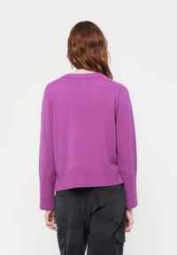 Pull en maille épaisse de couleur violet avec un ourlet et des poignets côtelés. L'arrière présente un col rond simple et des manches ajustées. Porté avec un pantalon sombre.