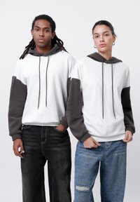 Sudadera gris claro con detalles en felpa gris oscuro, con cordón ajustable y un corte relajado. Lleva puesto unos pantalones de mezclilla negros y azules.