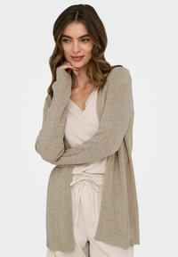 Cardigan beige en tricot à manches longues, à l'avant ouvert et avec des bords côtelés. Porté par-dessus un haut beige clair, mettant en valeur une coupe décontractée et une texture légère.