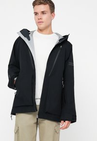 Noor noormees kannab musta lukuga jope koos kapuutsiga, mille all on valge T-särk ja beežid cargo-püksid, seistes ühtlase valge tausta ees.