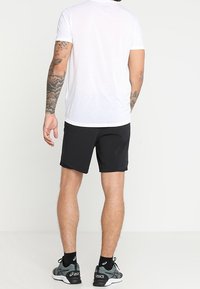 Black athletic shorts with a smooth texture, worn with a white crew-neck t-shirt. Model features visible tattoos and gray athletic shoes.